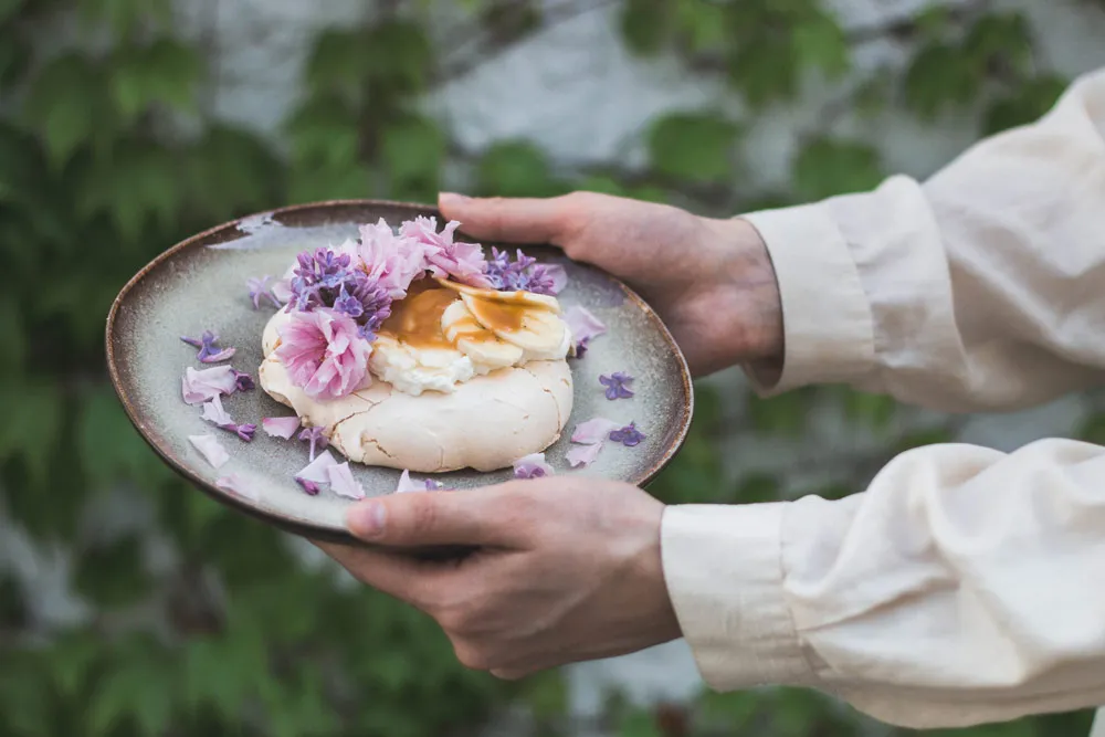 Mini pavlova s banány, karamelem a květy šeříku na keramickém talíři v dlaních u zdi s břečťanem