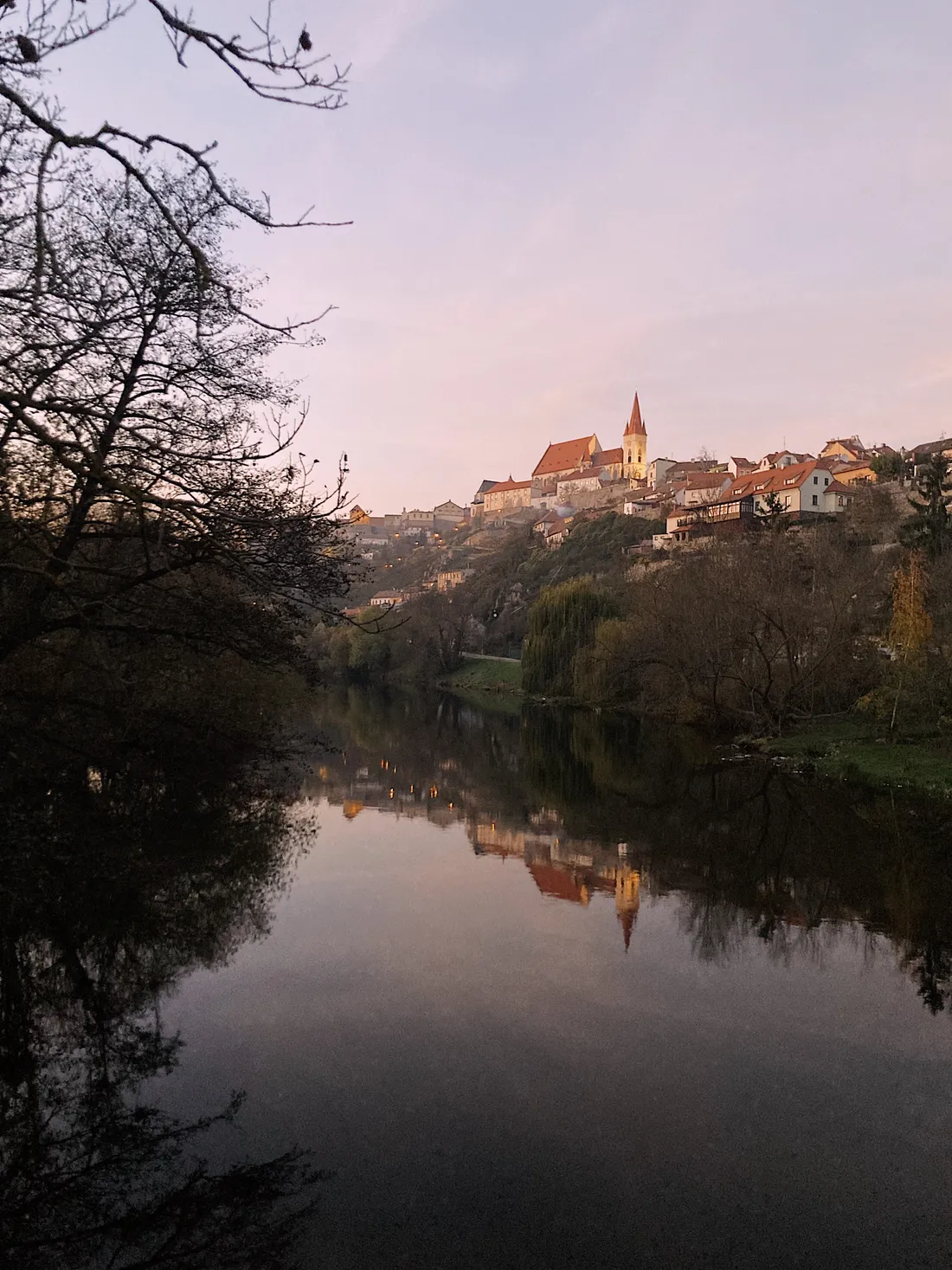 Historické město na kopci nad řekou při soumraku, kostel se špičatou věží se odráží ve vodě.