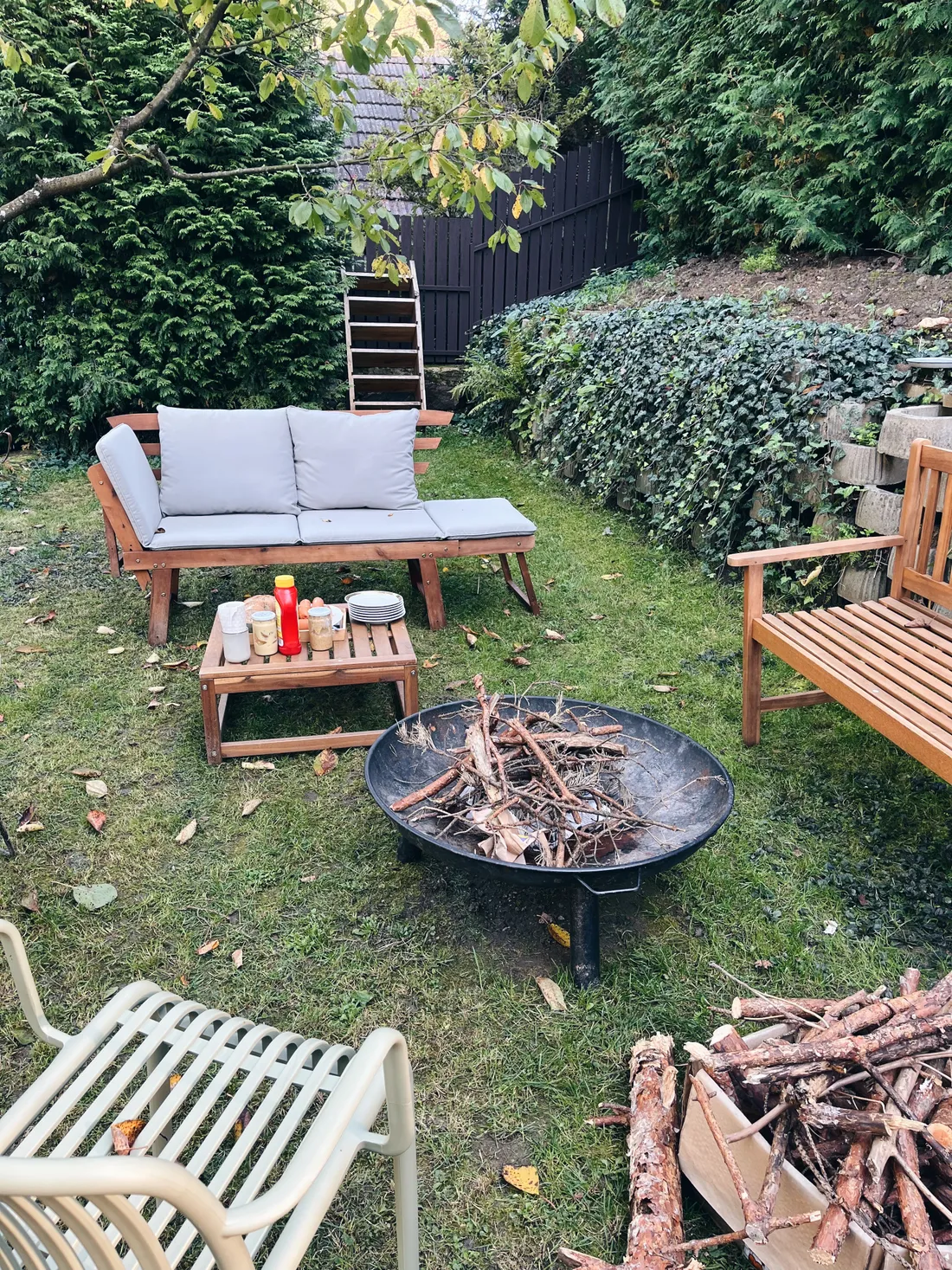 Zahradní posezení s dřevěnou pohovkou, stolkem s občerstvením a kovovým ohništěm na trávě.