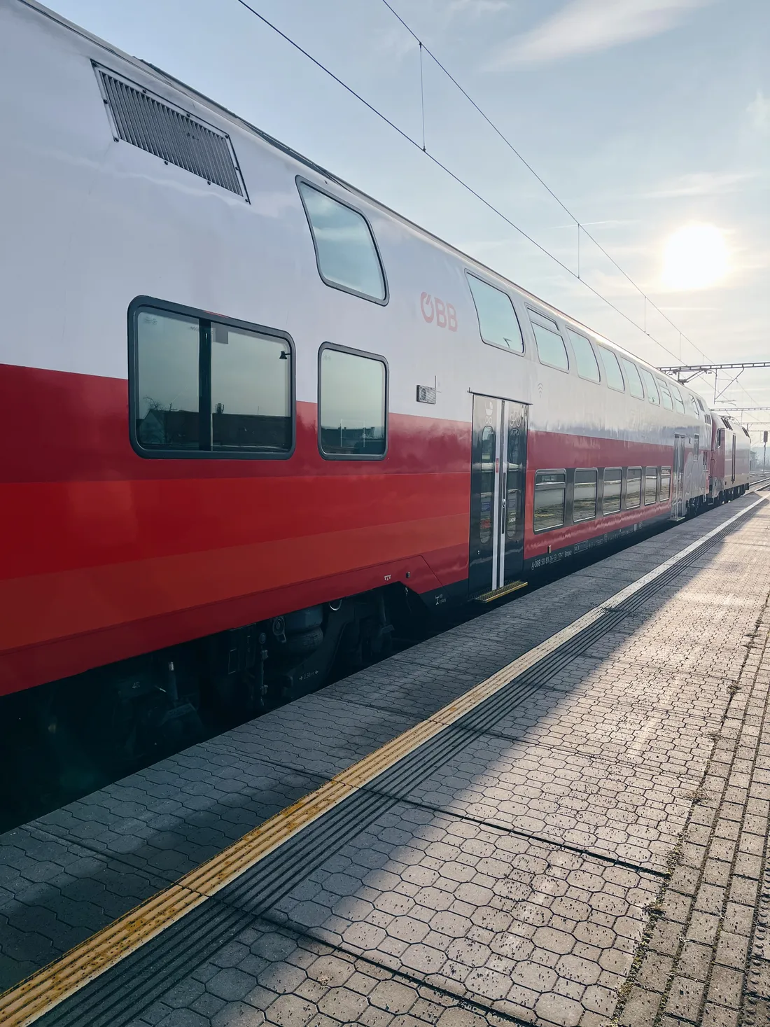 Červenobílý patrový vlak ÖBB na nástupišti zalitém sluncem s žlutou vodicí linií.