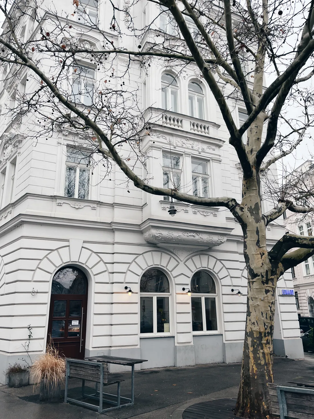 Bílá historická fasáda s arkádami, holý strom a dřevěná lavička na chodníku.