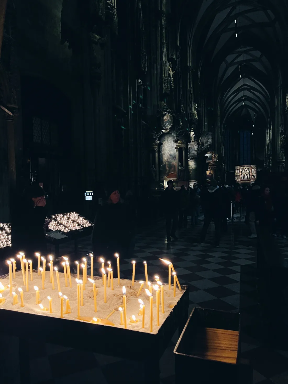Hořící svíčky v katedrále na kovovém stojanu, šachovnicová podlaha a tmavé klenby.