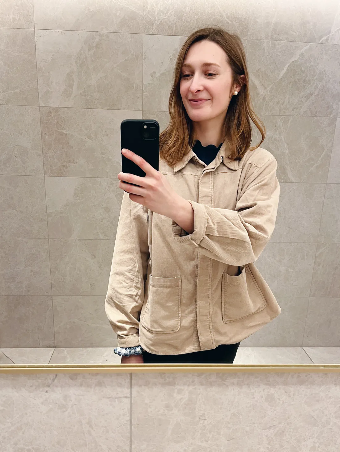 Žena v manšestrové bundě fotí selfie v zrcadle s mramorovou obkládačkou na pozadí.