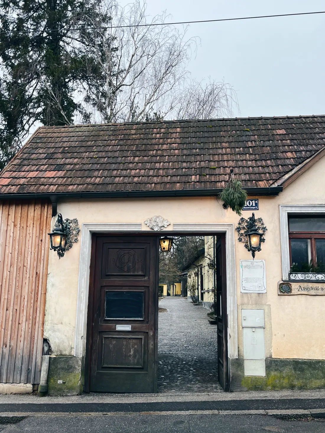 Vstup do heurigeru s otevřenou dřevěnou bránou, lucernami a jehličím nad vchodem.