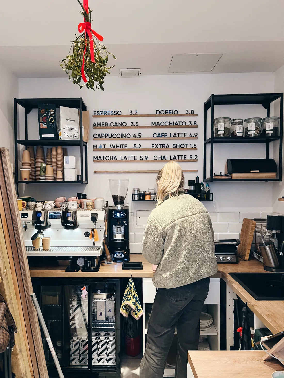 Baristka v kavárně u espresso stroje, dřevěné menu a jmelí visící ze stropu.