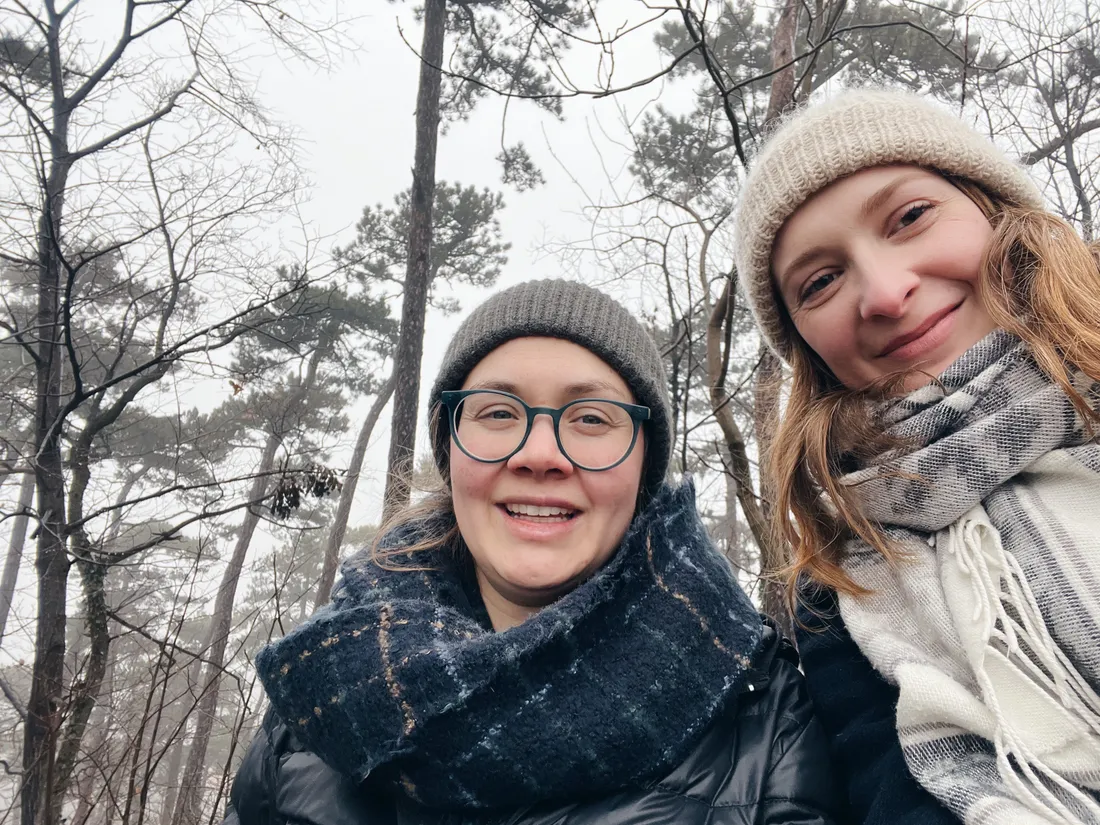 Dvě usmívající se ženy v čepicích a šálách na selfie v mlhavém lese.