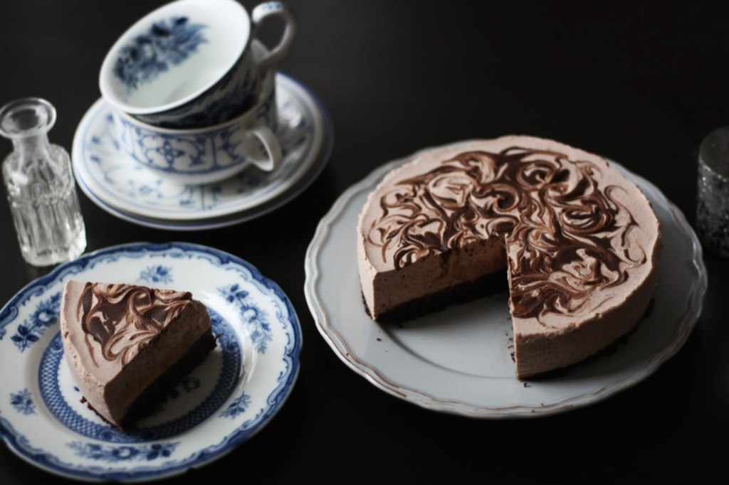 Čokoládový cheesecake s mramorováním, ukrojený dílek na modrobílém talíři a šálky v pozadí na tmavé ploše.