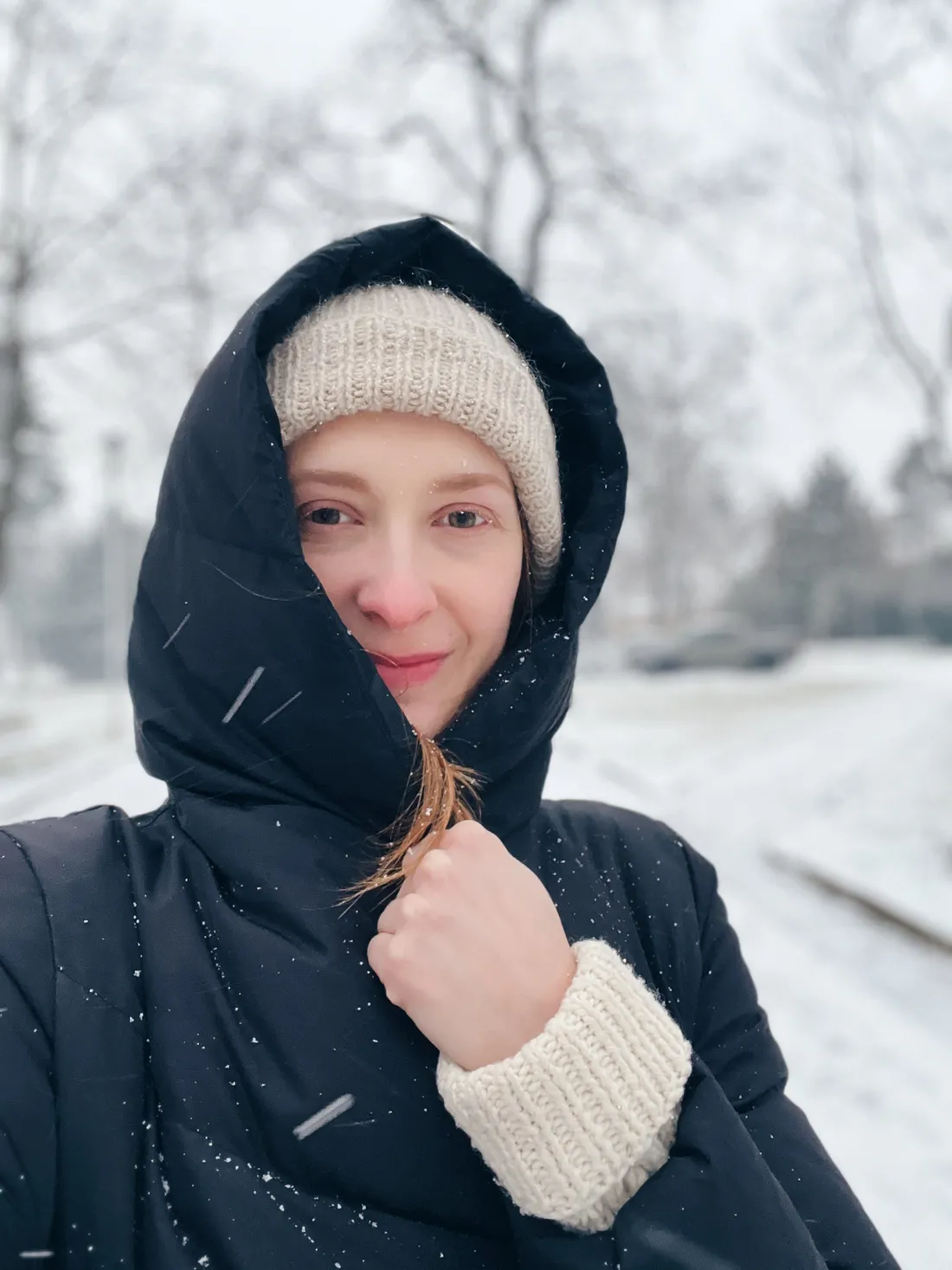 Selfie ženy ve sněhu v černé bundě a béžové čepici, drží pramínek vlasů.