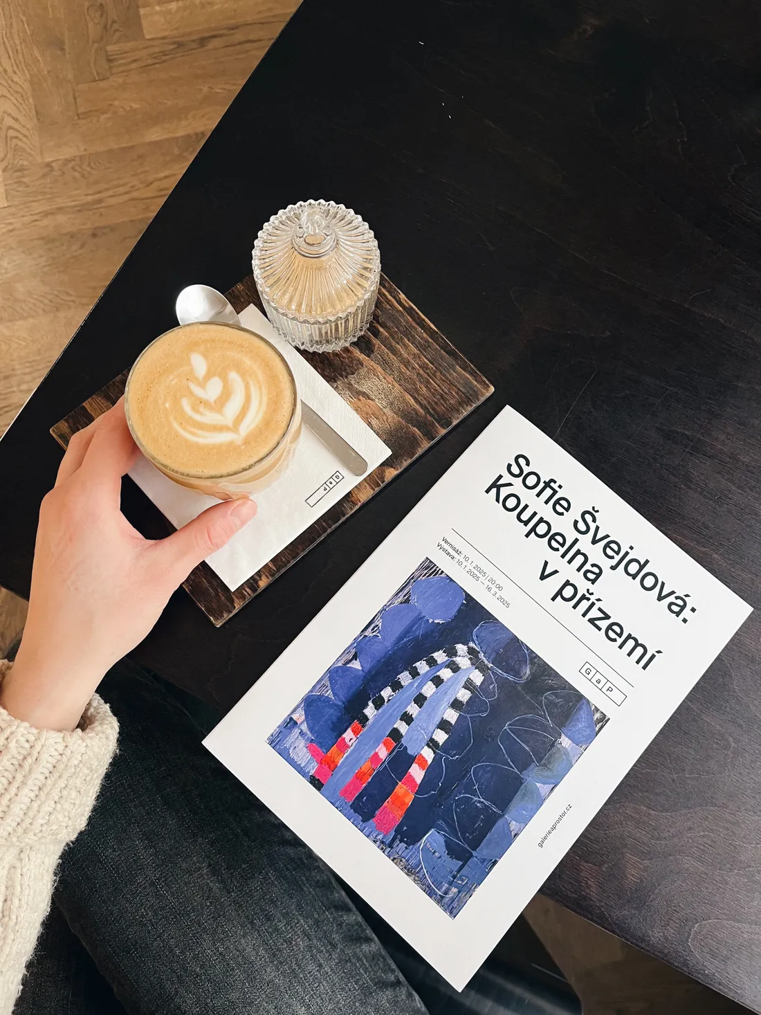 Latte s latte art a leták výstavy Sofie Švejdové na dřevěném podnose v kavárně.