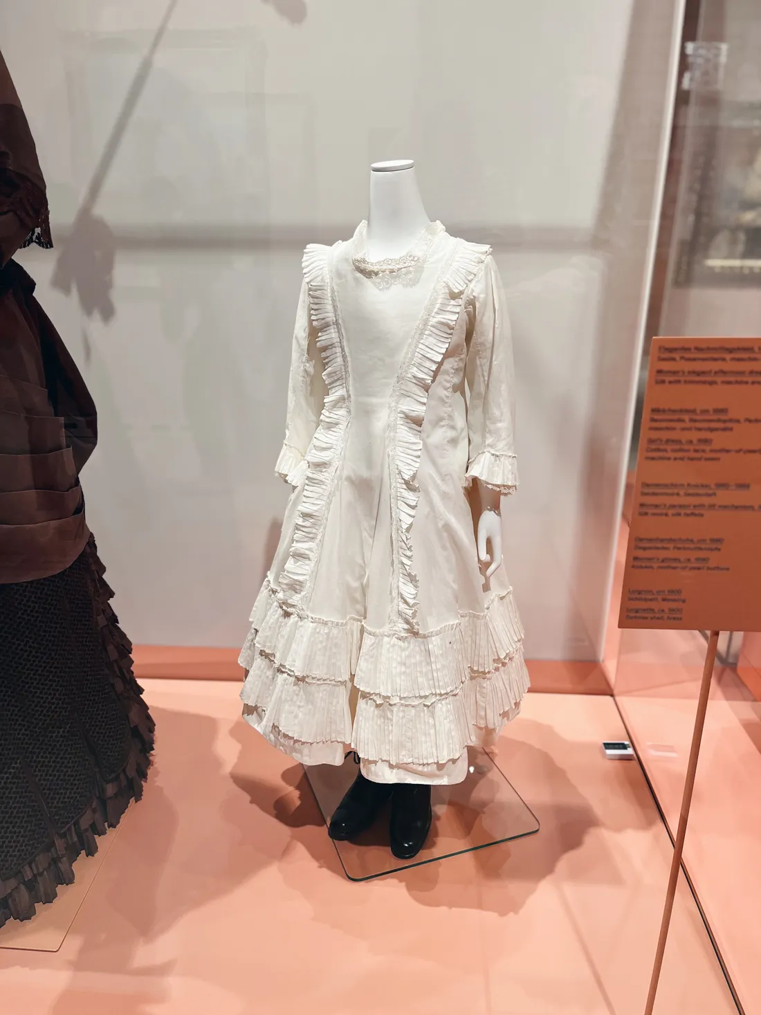 Historické bílé dětské šaty s volány na figuríně v muzejní expozici.
