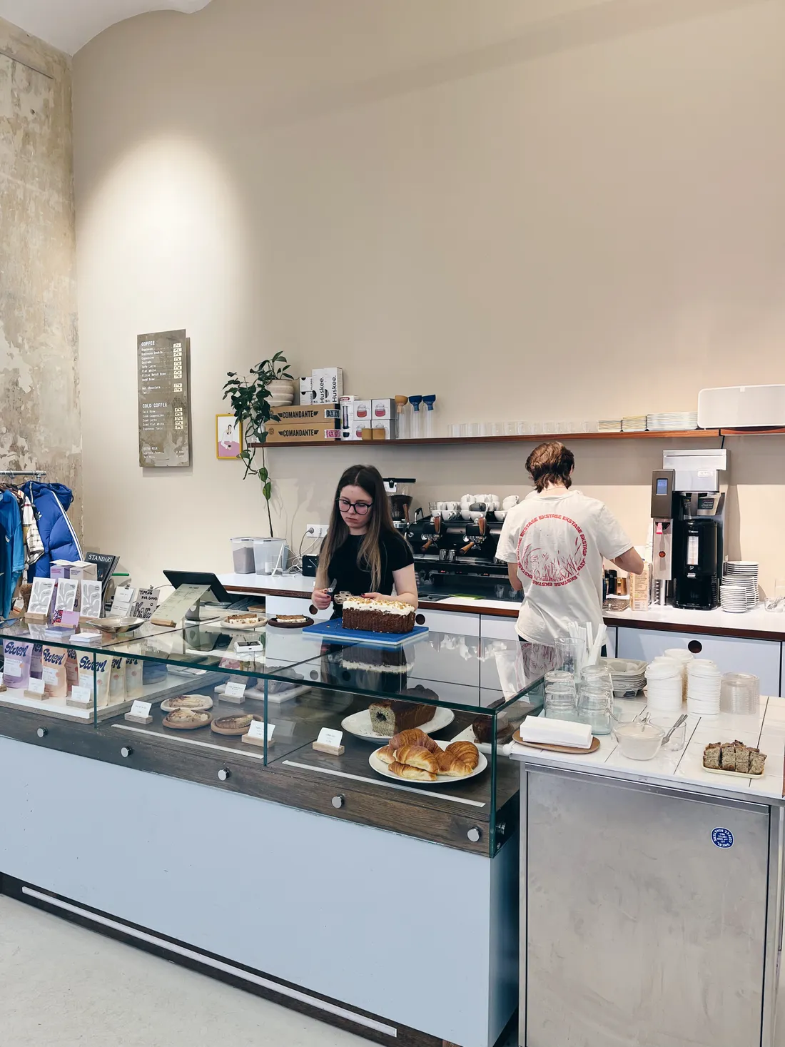 Interiér kavárny: baristka krájí dort za vitrínou s croissanty a zákusky.