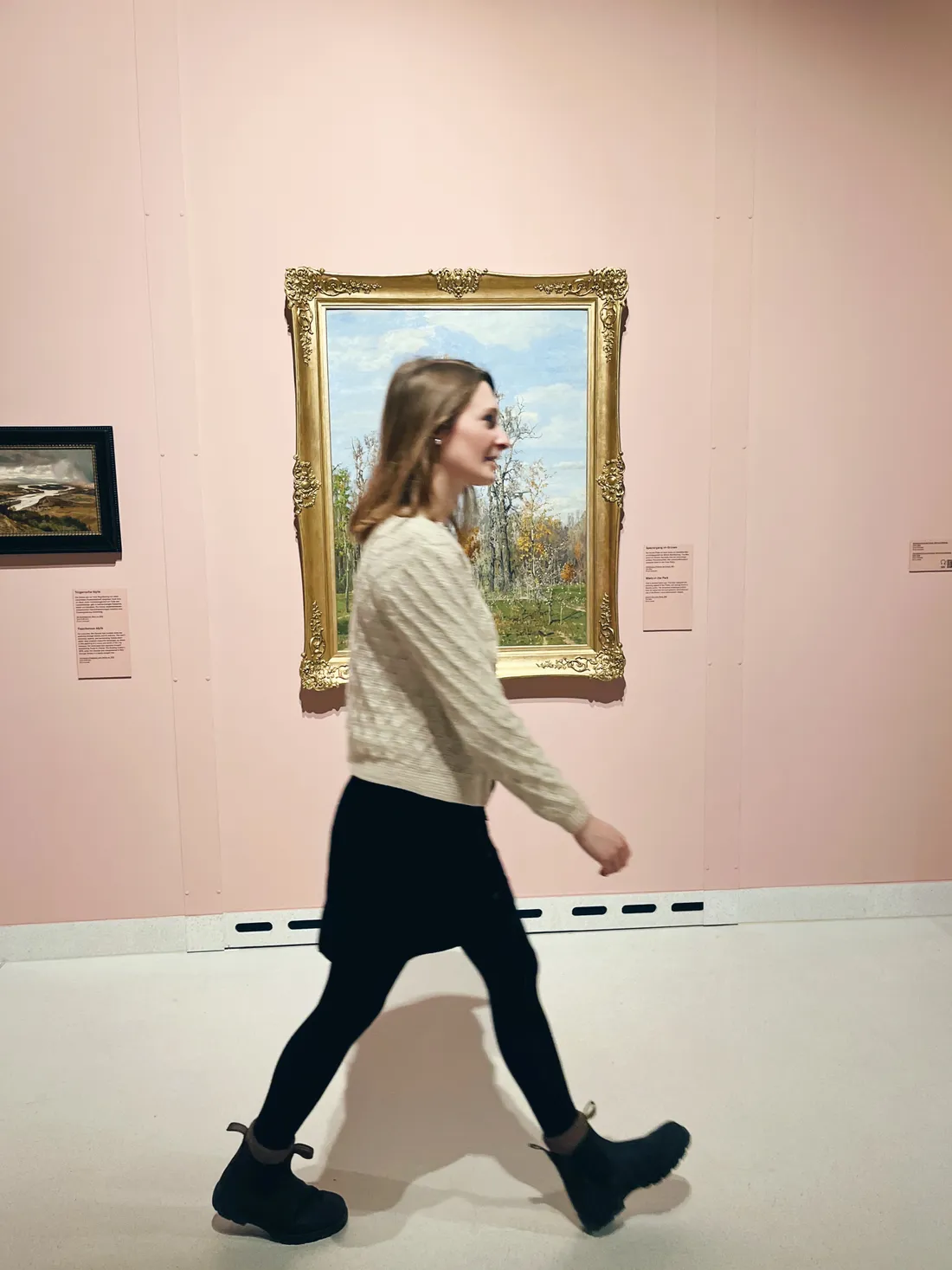Žena v černé sukni kráčí kolem obrazu v galerii s růžovou stěnou.