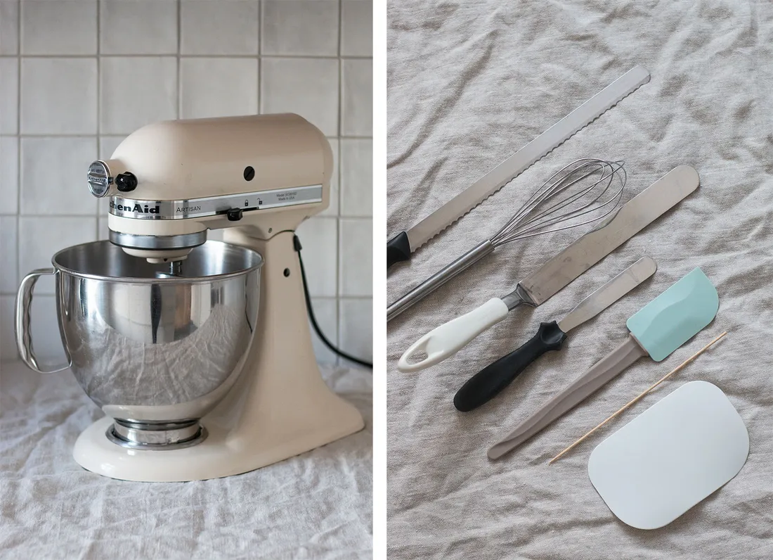 Krémový robot KitchenAid s nerezovou mísou na lněném ubrusu a sada pekařského náčiní na šedém plátně vedle sebe.