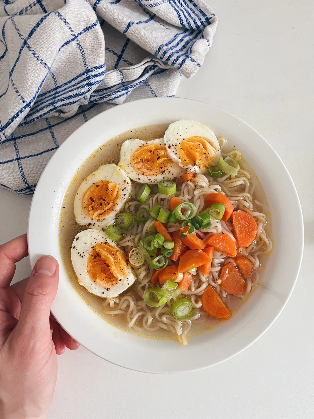 Miska ramen s čtyřmi vejci, mrkví a jarní cibulkou, ruka drží bílou misku.