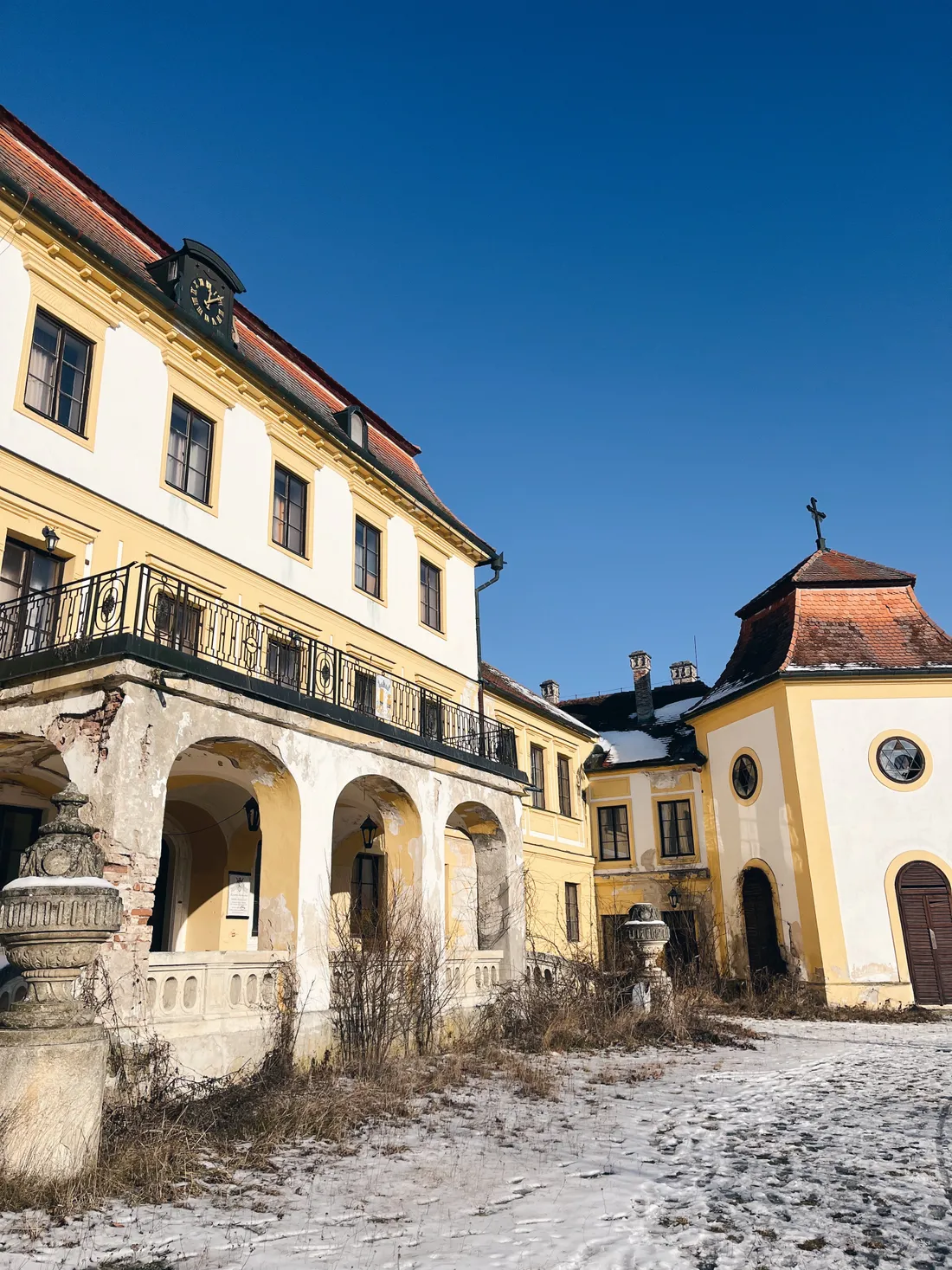 Žlutobílý zámek s arkádami a věžičkou na podzimním nádvoří se sněhem na zemi.
