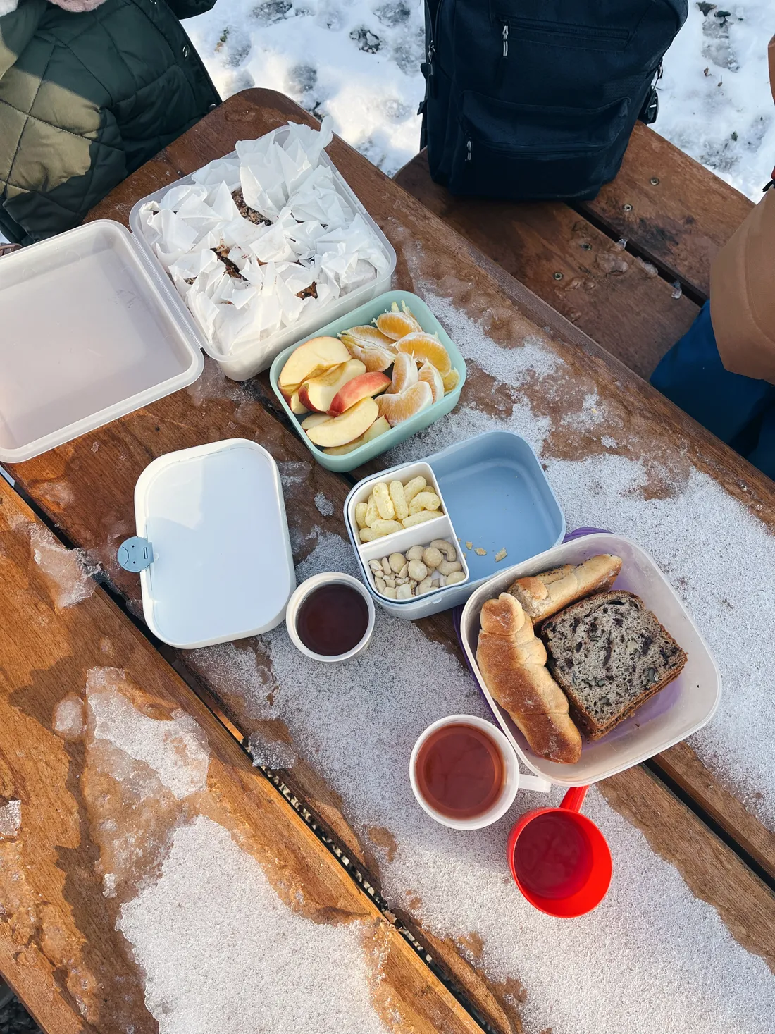 Zimní piknik na sněhu: krabice se svačinou, croissanty, ovoce a horké nápoje.