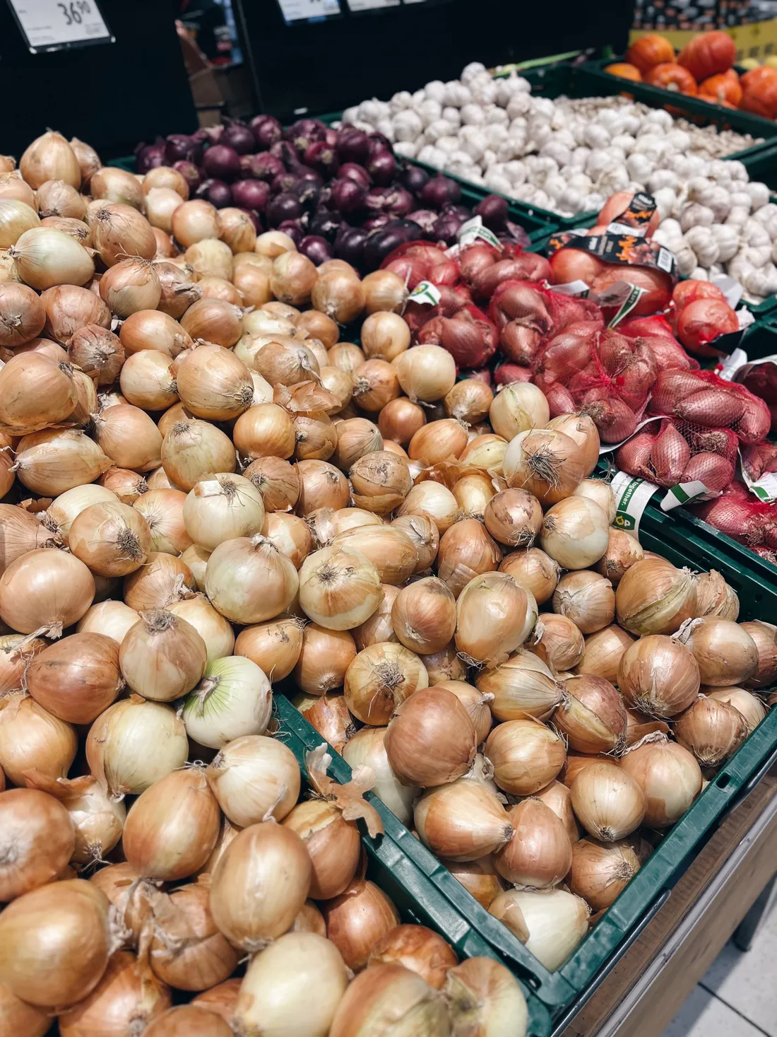 Zelené bedny v obchodě plné žluté, červené cibule, šalotky v síťkách a bílého česneku.