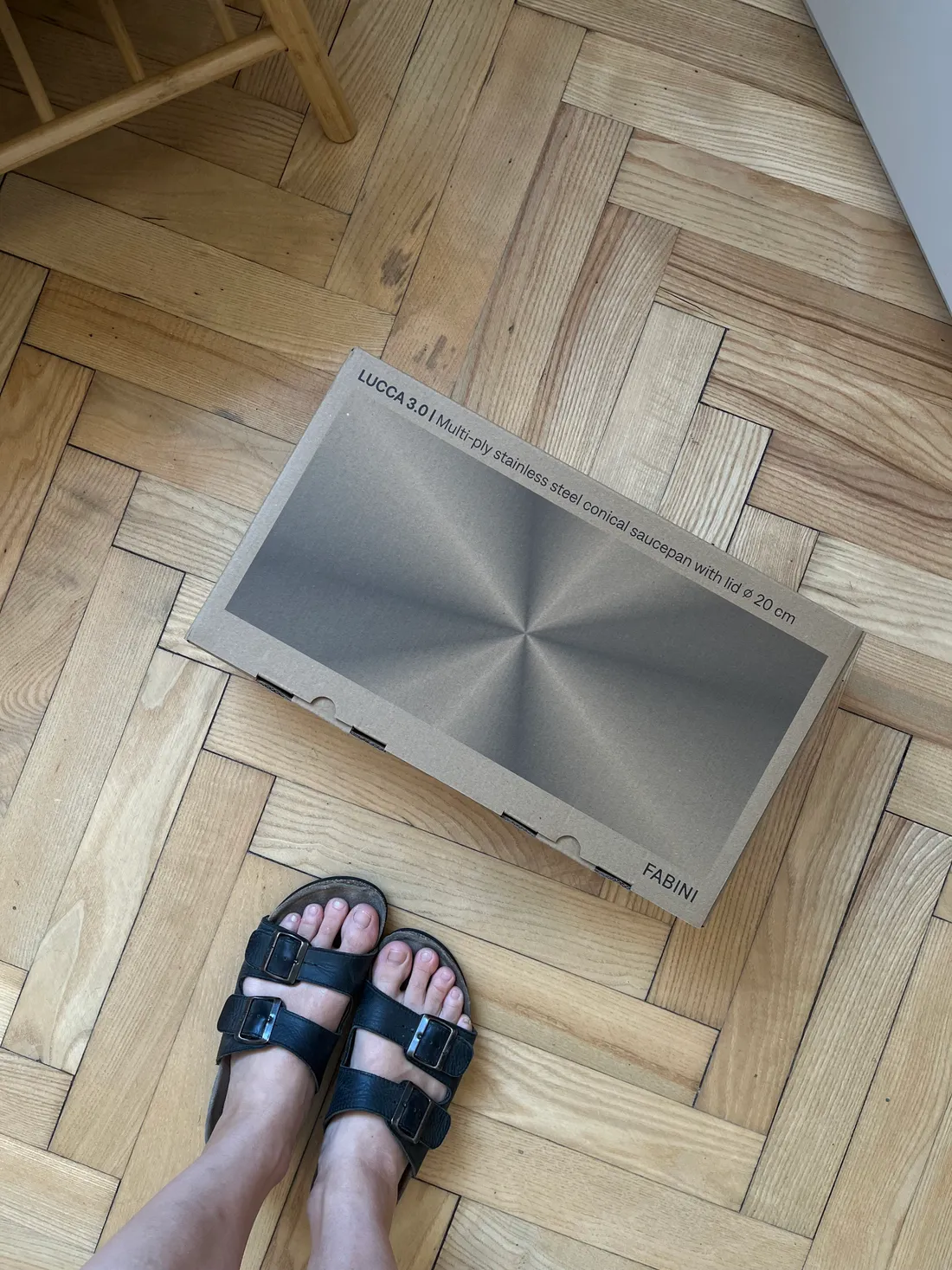 Krabice s nerezovým kastrolem Fabini Lucca 3,0 l na parketách, v popředí nohy v páskových sandálech.