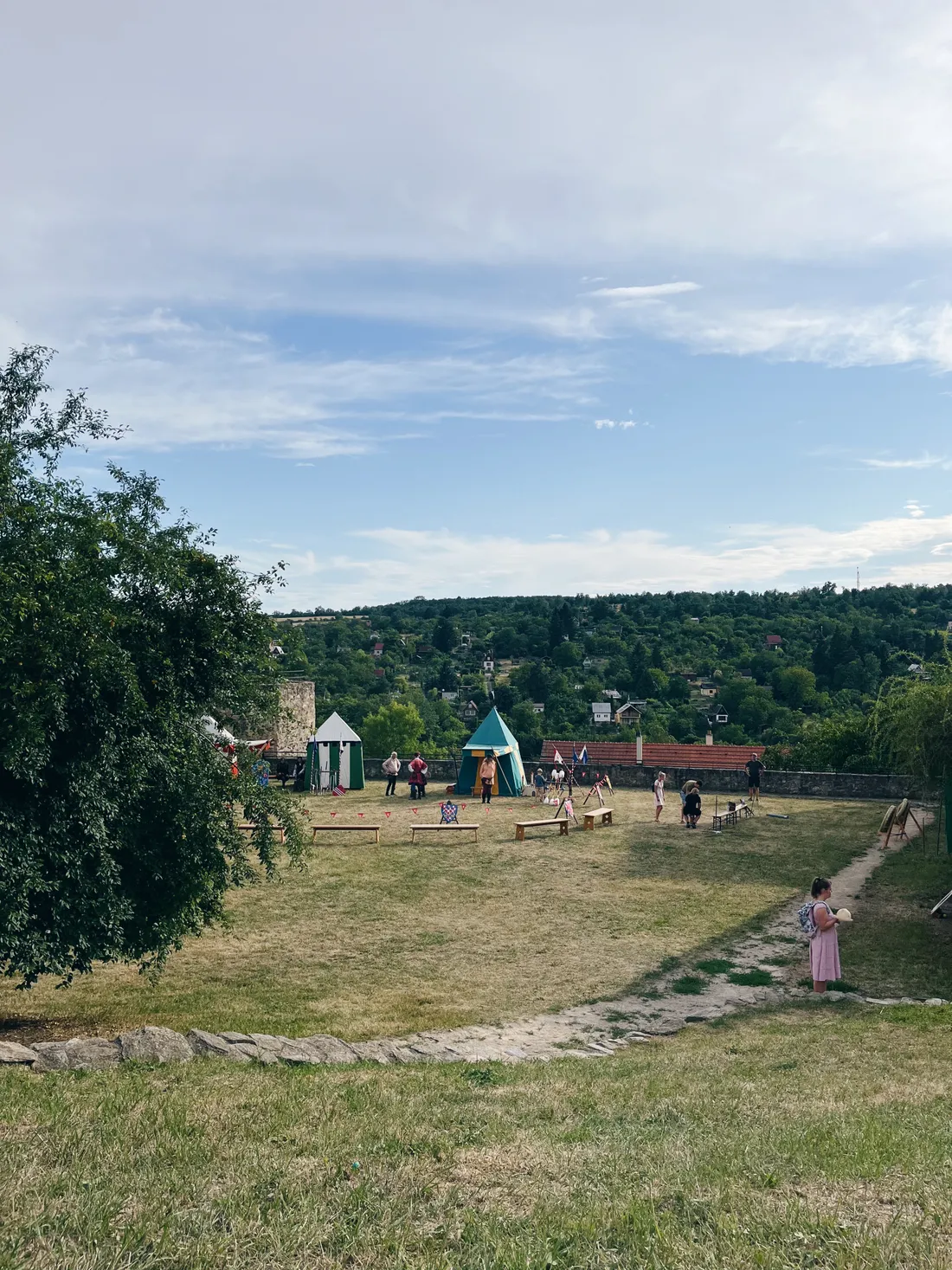 Travnaté prostranství s historickými stany a kostýmy pod zalesněným kopcem.