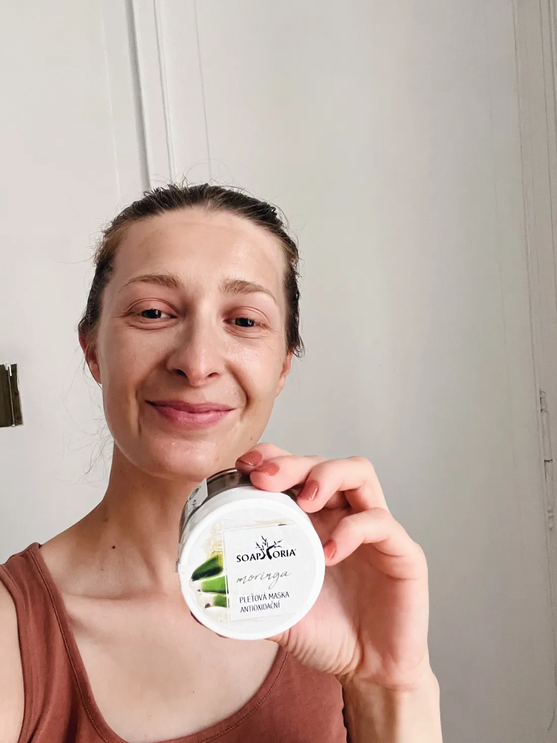 Usmívající se žena drží bílou sklenici pleťové masky Soaphoria Moringa.