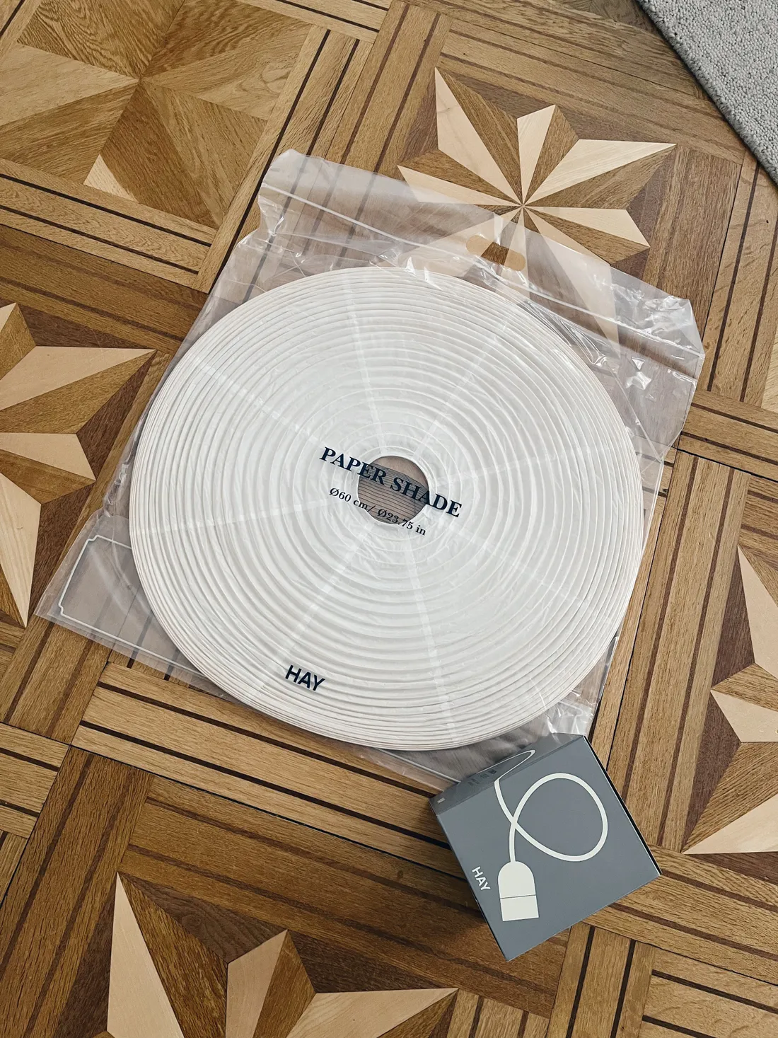 Složený bílý papírový lampion HAY Paper Shade Ø60 cm a malá šedá krabička na parketách.