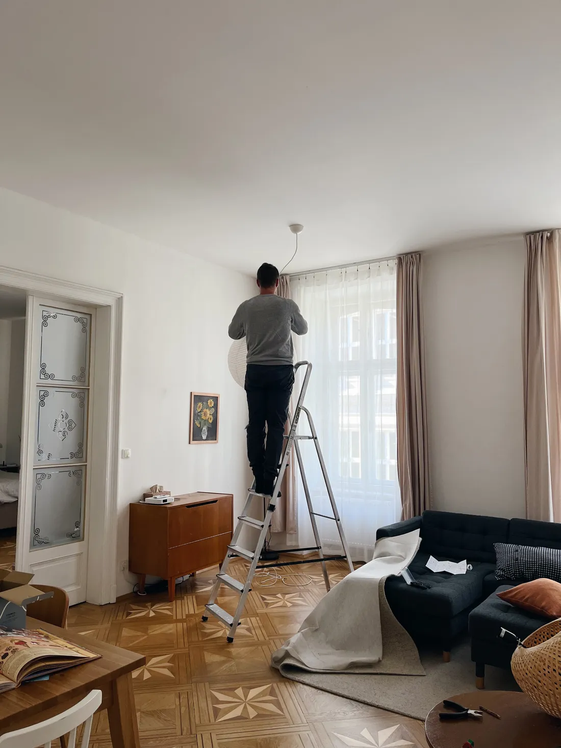 Muž na žebříku montuje bílou papírovou lampu v obýváku s parketami a šedou pohovkou.
