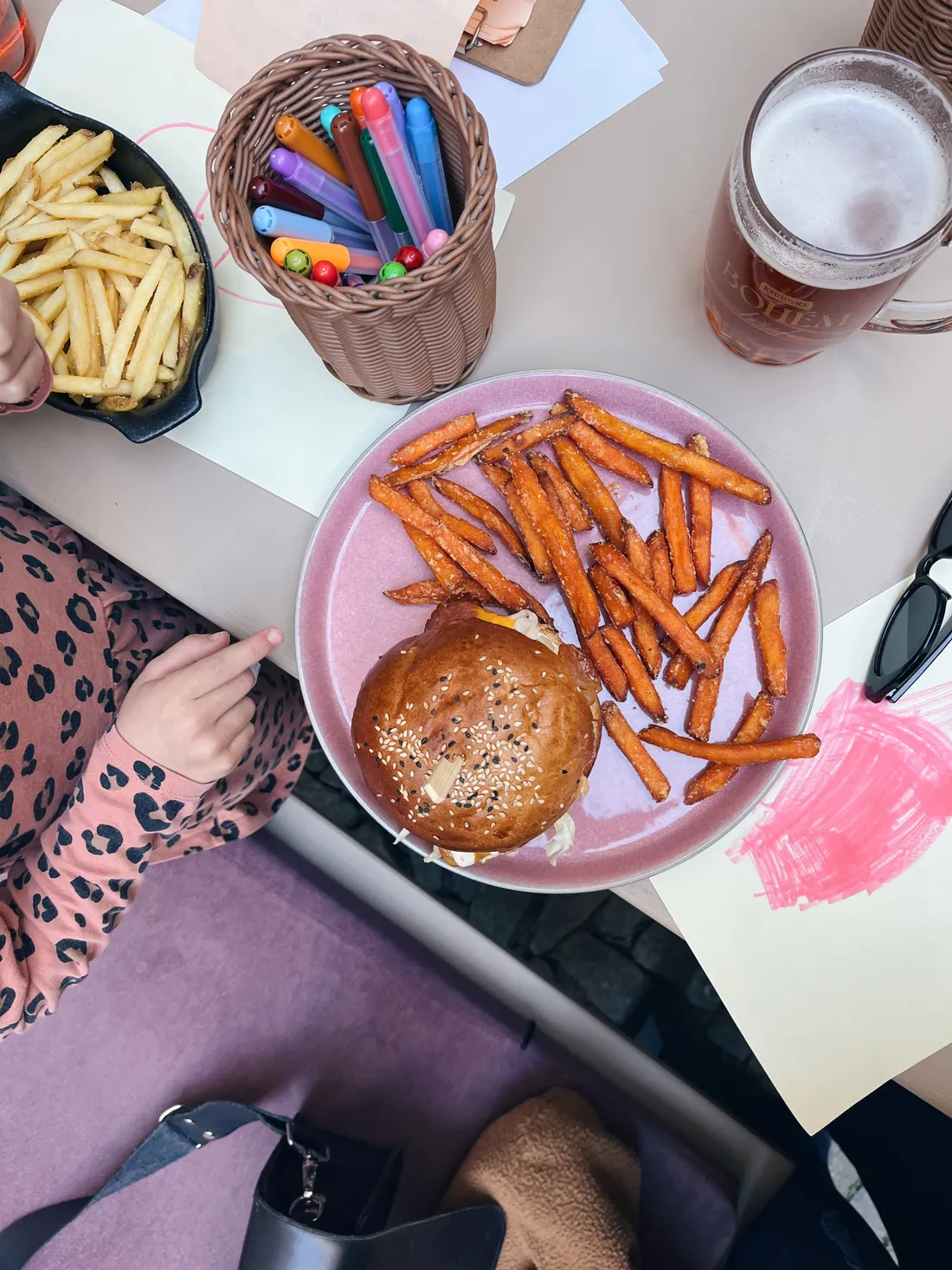 Burger se sladkými bramborami na růžovém talíři, pivo a košík fixů, dětská ruka ukazuje na jídlo.