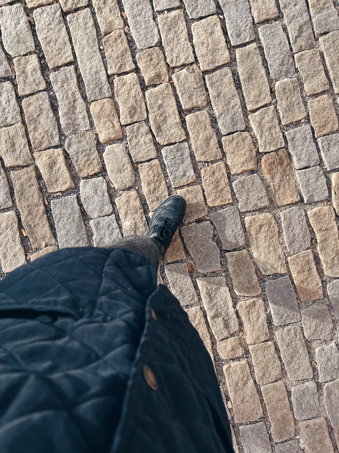 Černá krajková bota na barevné kamenné dlažbě, pohled z první osoby.