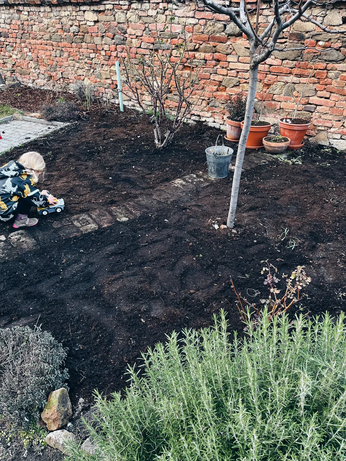 Dítě hraje s bagrem na cihlové cestě v záhonu s čerstvou hlínou u kamenné zdi.