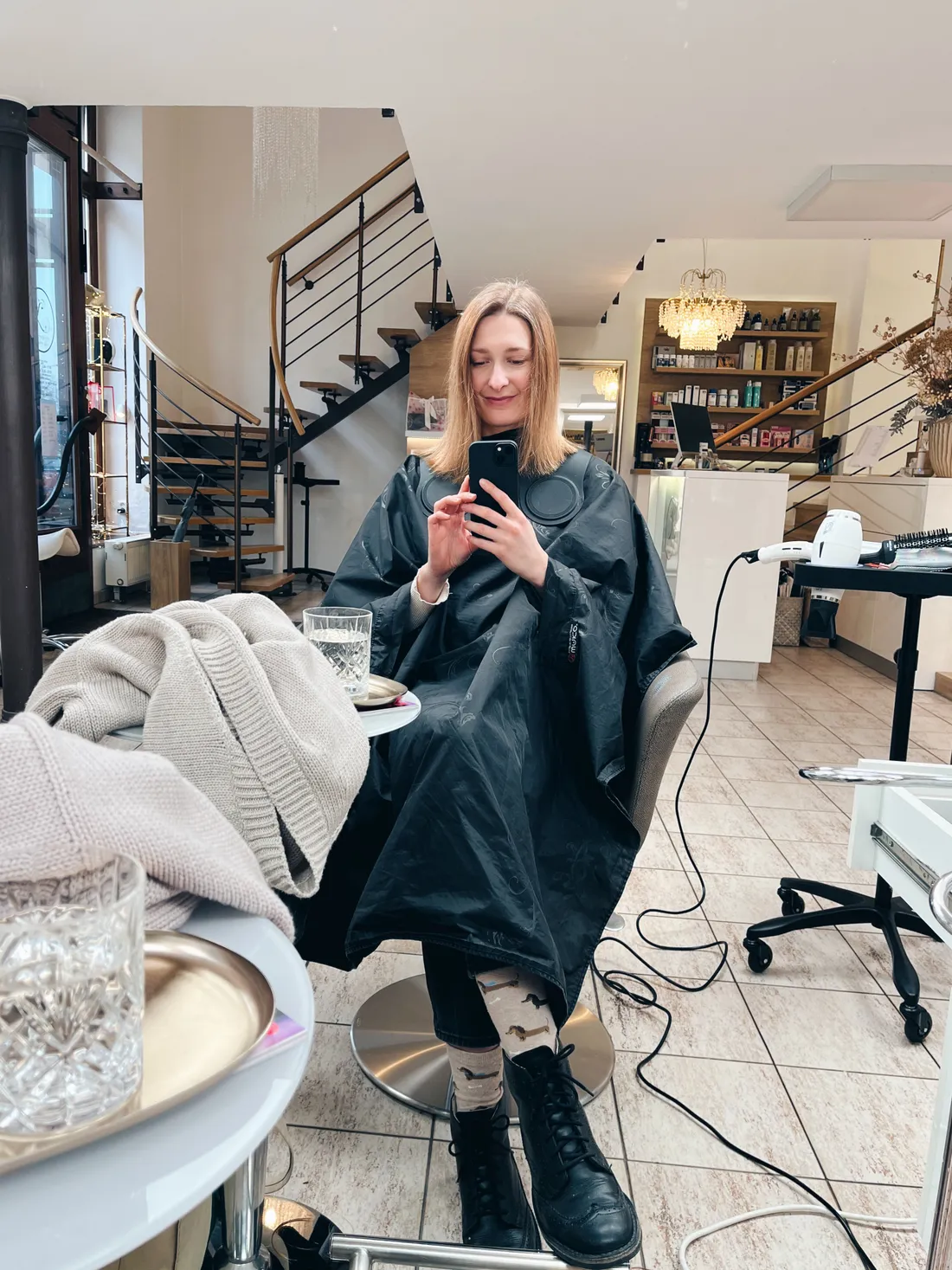 Žena v černém kadeřnickém plášti fotí se v zrcadle v moderním salonu se spirálovým schodištěm.
