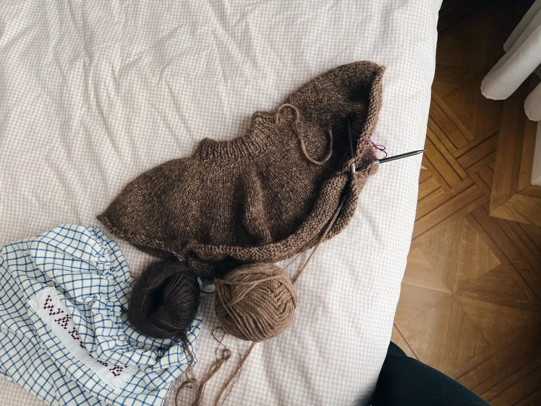 Rozpletený hnědý svetr na jehlících na posteli vedle klubka tmavé a světle hnědé příze v modro-bílém pytlíku.