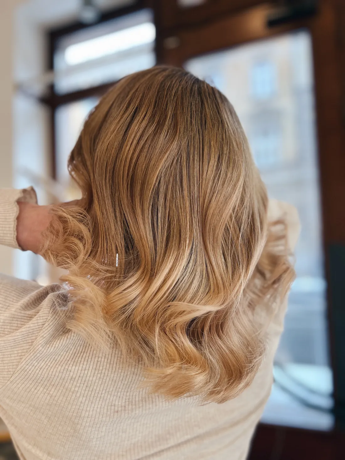 Záběr na medově blond vlasy ve vlnách, ruce u krku, světlý svetr u okna.