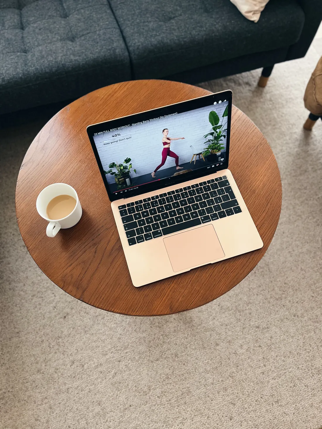 Růžový notebook s fitness videem na kulatém stolku vedle bílého šálku kávy a šedé pohovky.