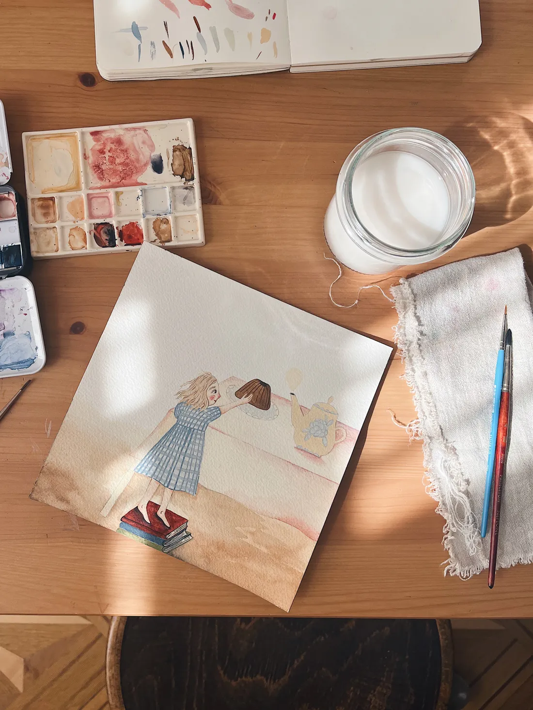 Na dřevěném stole akvarelová ilustrace dívky na knihách u bábovky, kolem paleta, štětce a sklenice s vodou.