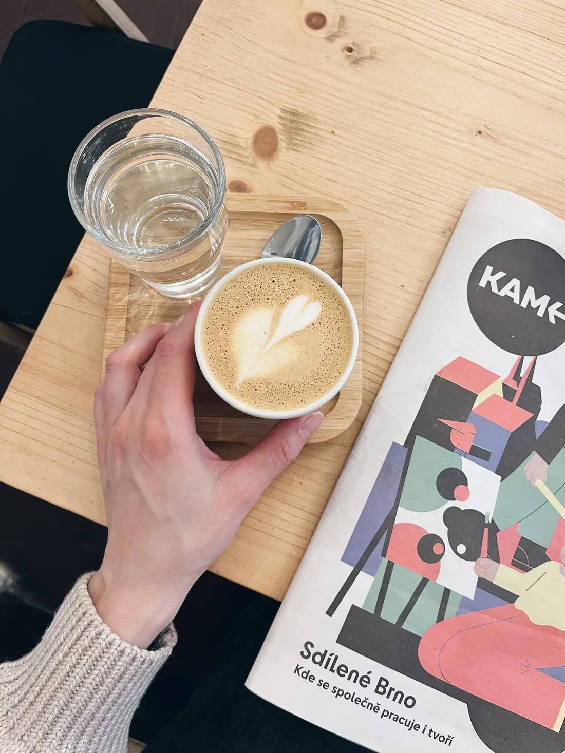 Ruka drží bílý šálek cappuccina s latte art srdcem na dřevěném podnosu vedle sklenice vody a časopisu KAM.