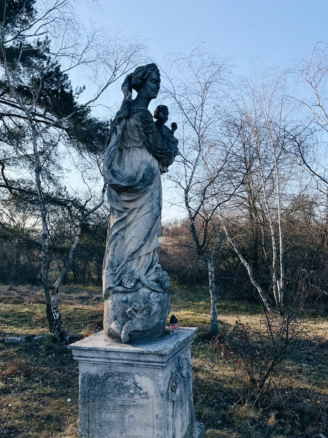 Zvětralá kamenná socha ženy s dítětem na podstavci mezi břízami v trávě pod jasnou oblohou.