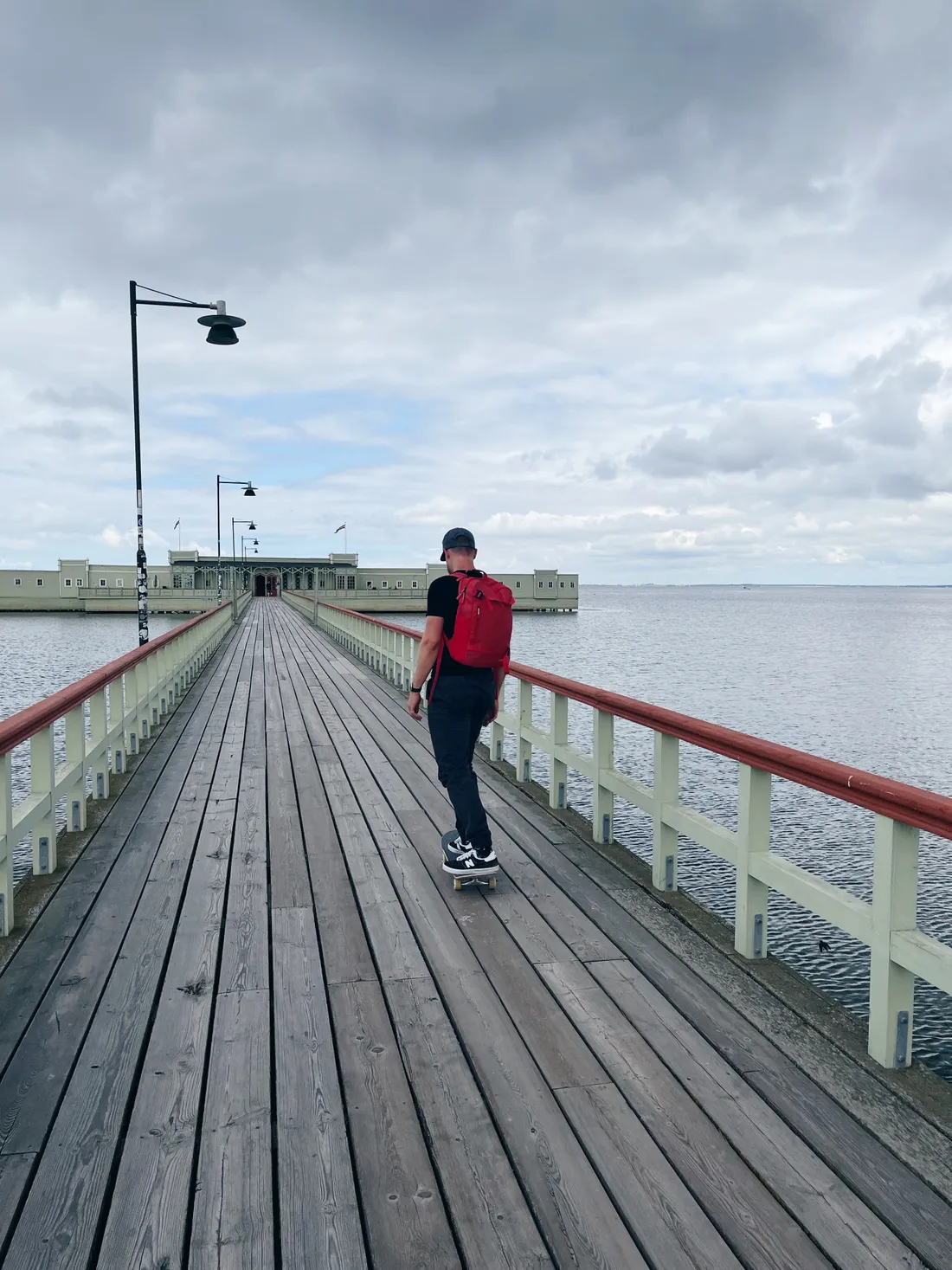 Muž na skateboardu s červeným batohem na dřevěném molu u moře pod šedou oblohou.