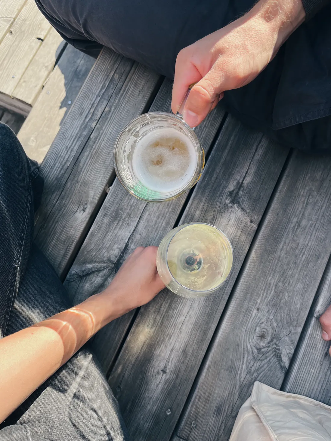 Dva lidé sedí na dřevěné terase, jeden drží pivo, druhý bílé víno ve slunečním světle.