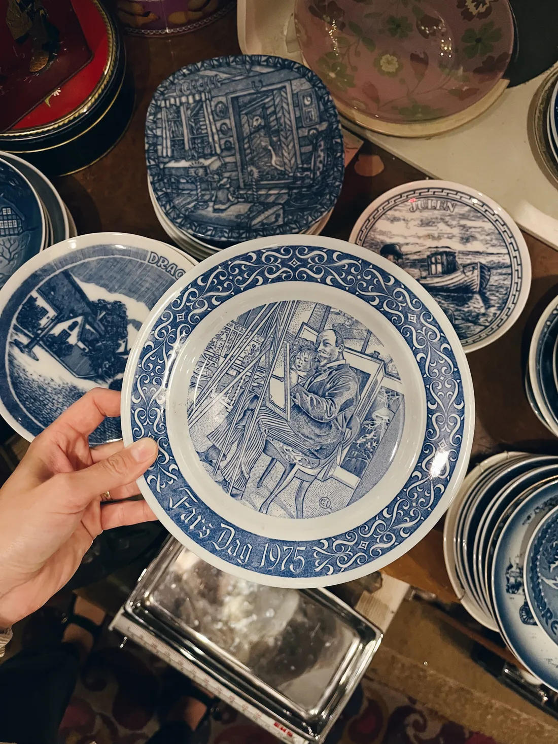 Ruka drží modrobílý porcelánový talíř s ilustrací malíře a dítěte, v pozadí další dekorativní talíře.