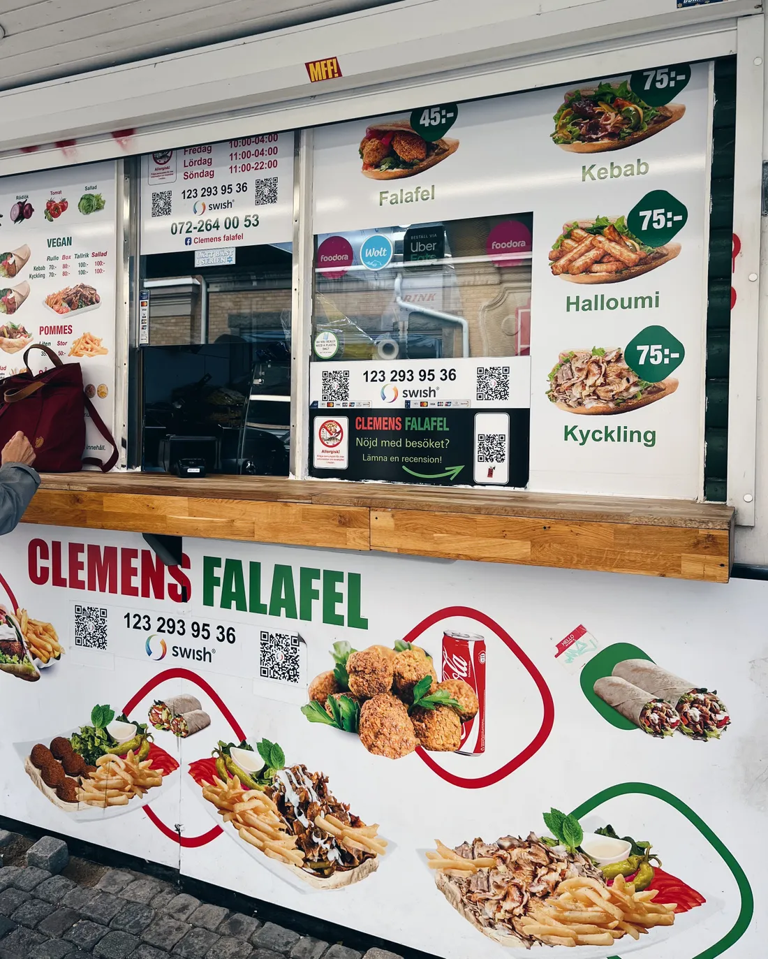 Stánek Clemens Falafel s menu falafelu, kebabu a halloumi a fotkami jídel na dřevěné výloze.