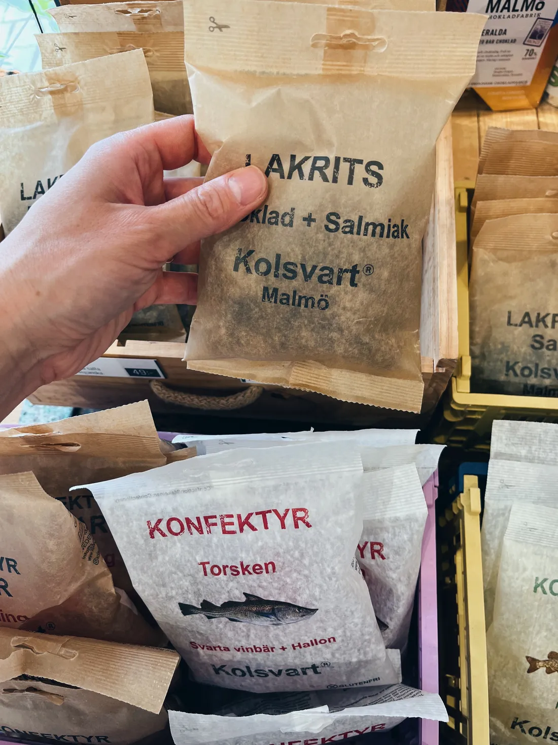 Ruka drží papírový sáček švédské lékořicové sladkosti Kolsvart v obchodě s dalšími baleními v přepravkách.