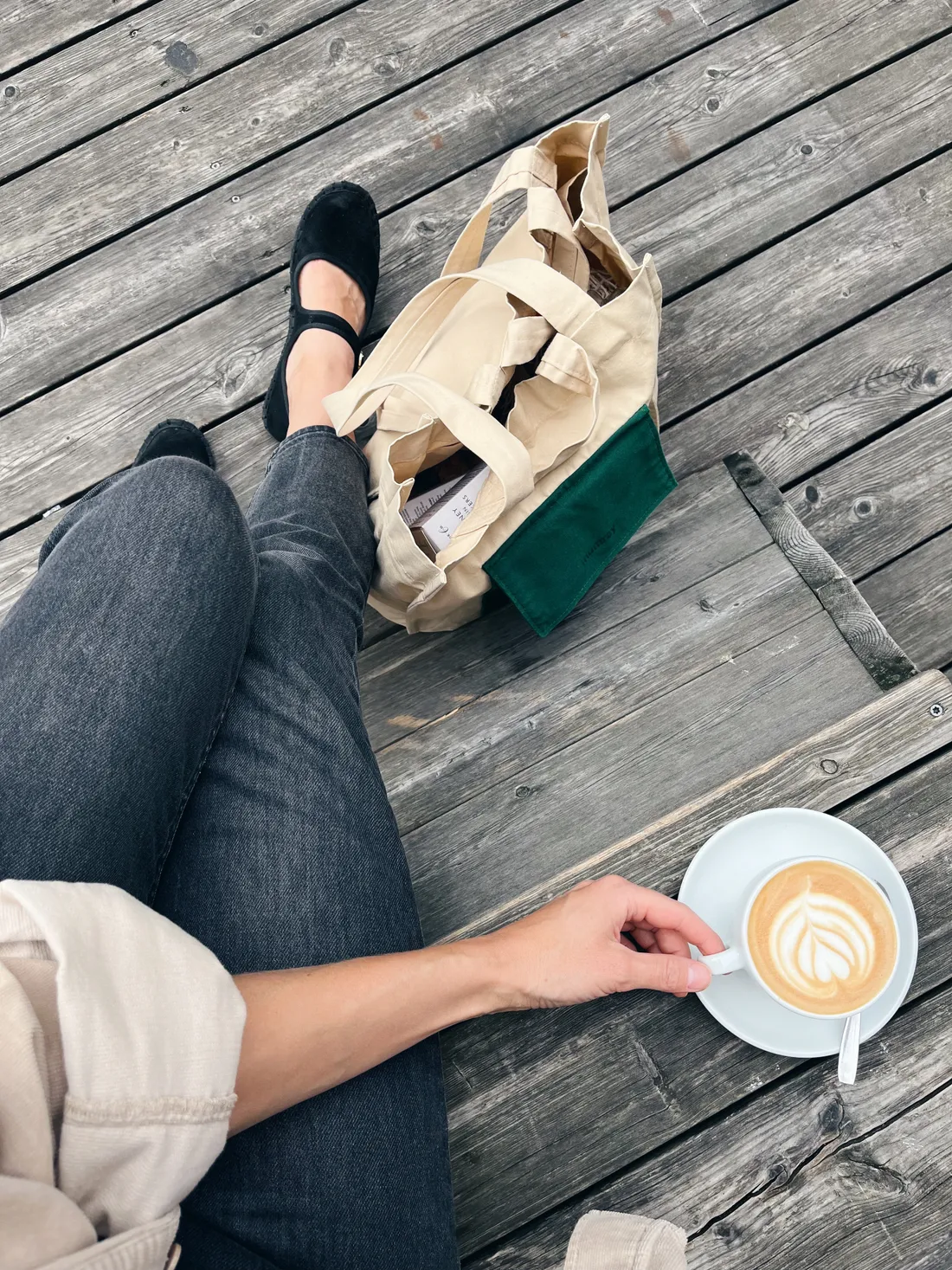 Ruka drží šálek kávy s latte art na dřevěné terase, vedle plátěná taška se zelenou kapsou.