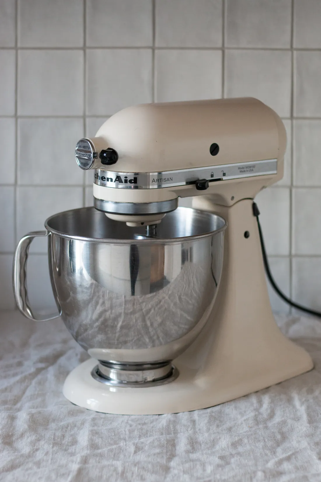 Krémový kuchyňský robot KitchenAid Artisan s nerezovou mísou na bílém ubrusu u kachličkované stěny.