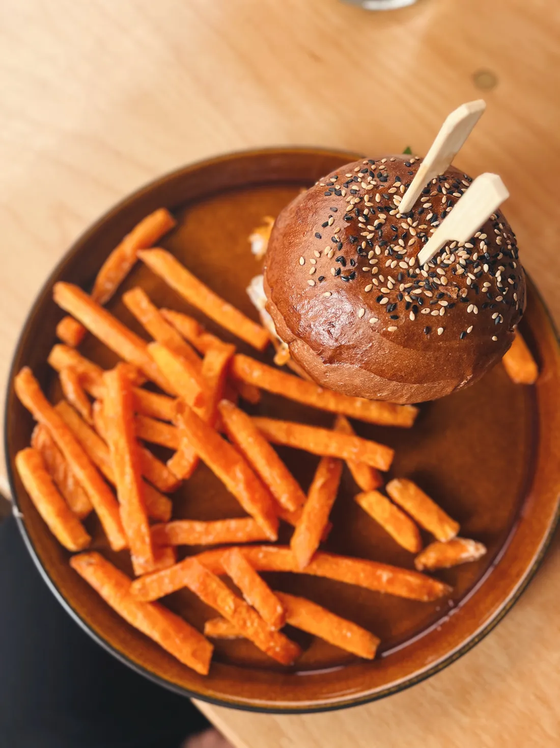 Burger se sezamovou houskou a hranolky ze sladké brambory na hnědém keramickém talíři na dřevěném stole.