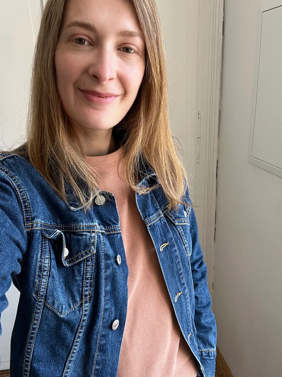 Žena v džínové bundě a broskvovém svetru se jemně usmívá na selfie u bílé stěny.