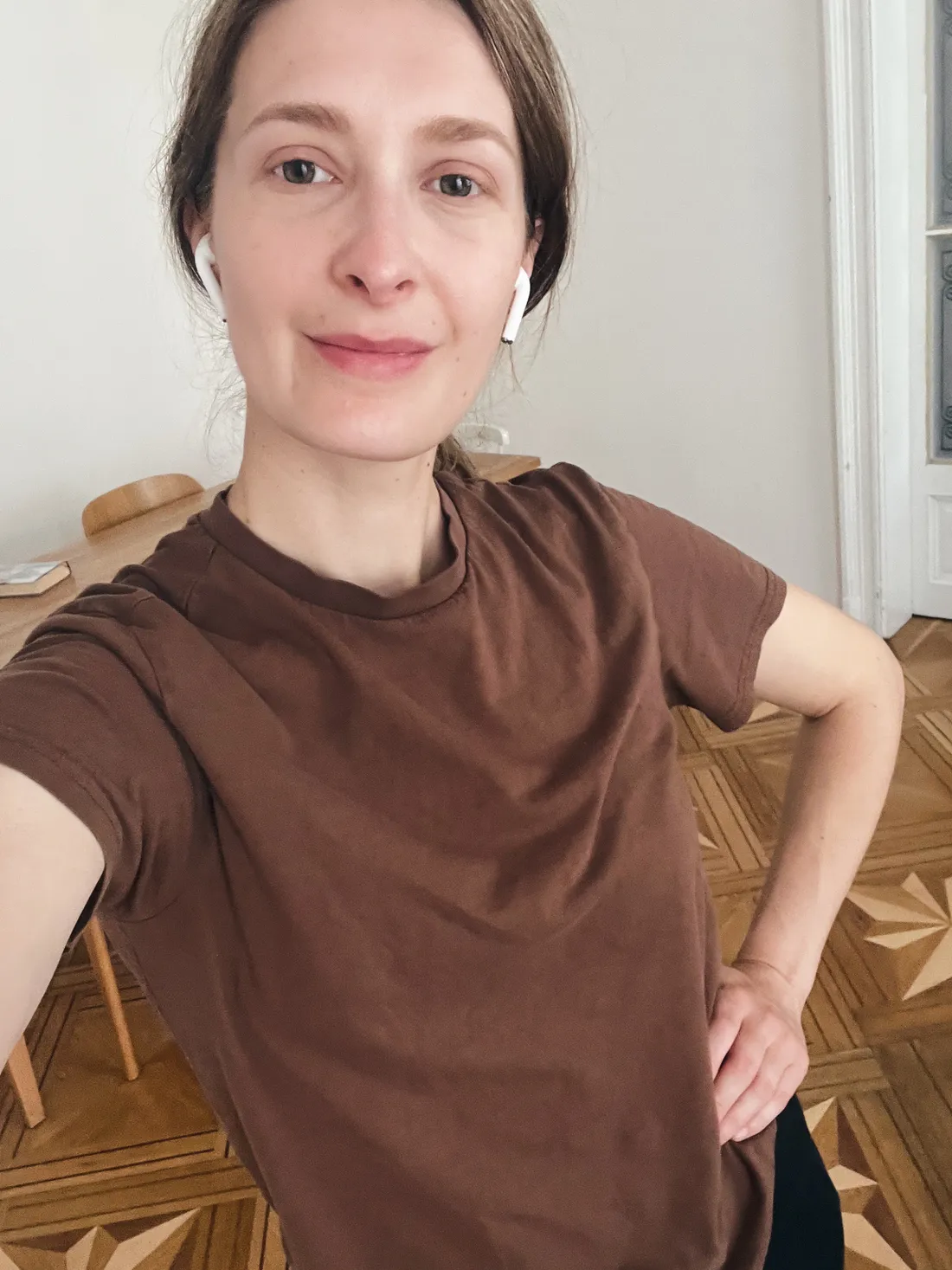 Selfie ženy s hnědými vlasy v béžovém triku a bílých sluchátkách v místnosti s parketami.