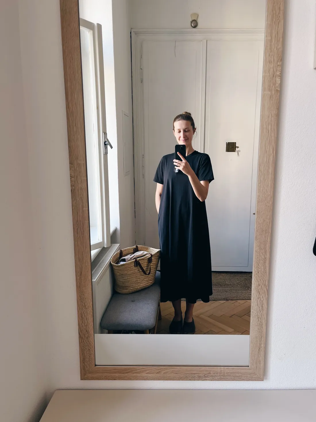 Žena v černých šatech v zrcadle s proutěnou taškou na lavici u okna.