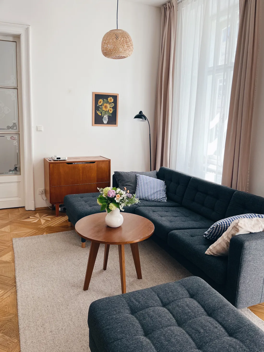 Obývací pokoj s tmavě šedou pohovkou, dřevěným stolkem, kyticí a proutěným lustrem u okna.
