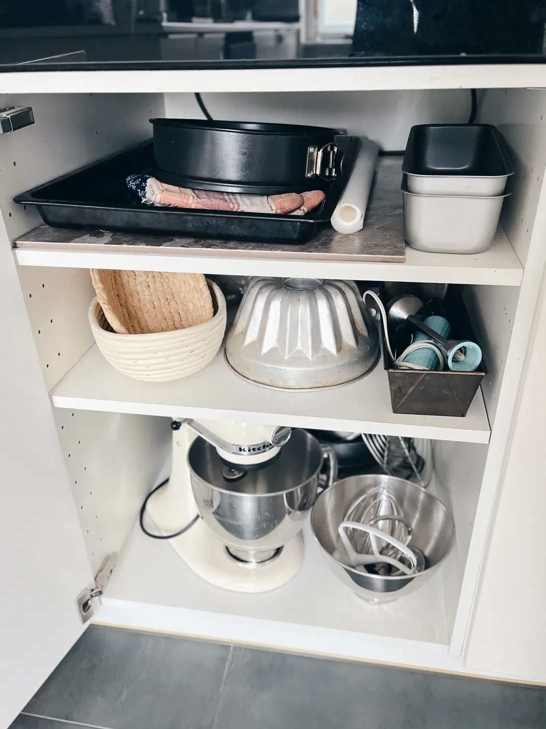Otevřená bílá skříňka s formami, proutěnými ošatkami, mixérem KitchenAid a válečkem na šedé dlažbě.