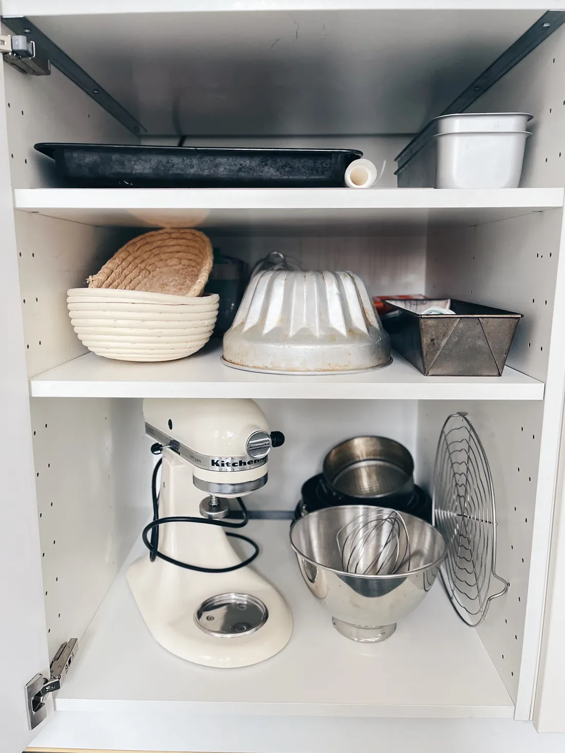 Otevřená bílá skříňka s robotem KitchenAid, nerezovými mísami, ošatkami, formami a chladicí mřížkou.